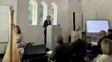 Auf dem Foto ist die NRW-Ministerin und stellv. Landesvorsitzende des Volksbundes, Dorothee Feller, bei Ihrem Vortrag in der Kapelle des evangelischen Friedhofs in Recklinghausen zu sehen. Links neben ihr sieht man eine Harfe-Spielerin, die die Veranstaltung musikalisch begleitet. Rechts neben der Ministerin sieht man einen großen Aufsteller inkl. Titel der Gedenkveranstaltung „Wir sind die Eingeborenen von Trizonesien“ - Der 8. Mai 1945 in Recklinghausen und Europa. Ein Tag der Befreiung?